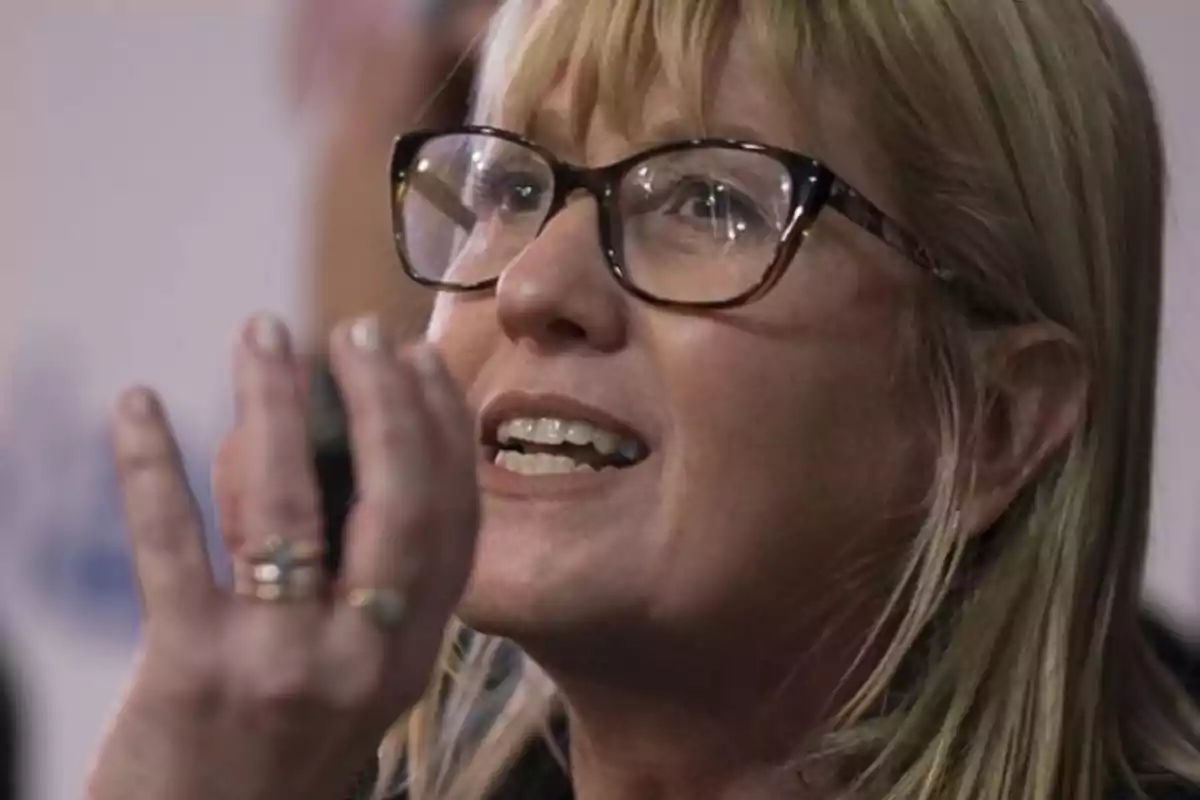 Blonde woman with large glasses talking and gesturing with her hand in front of her face
