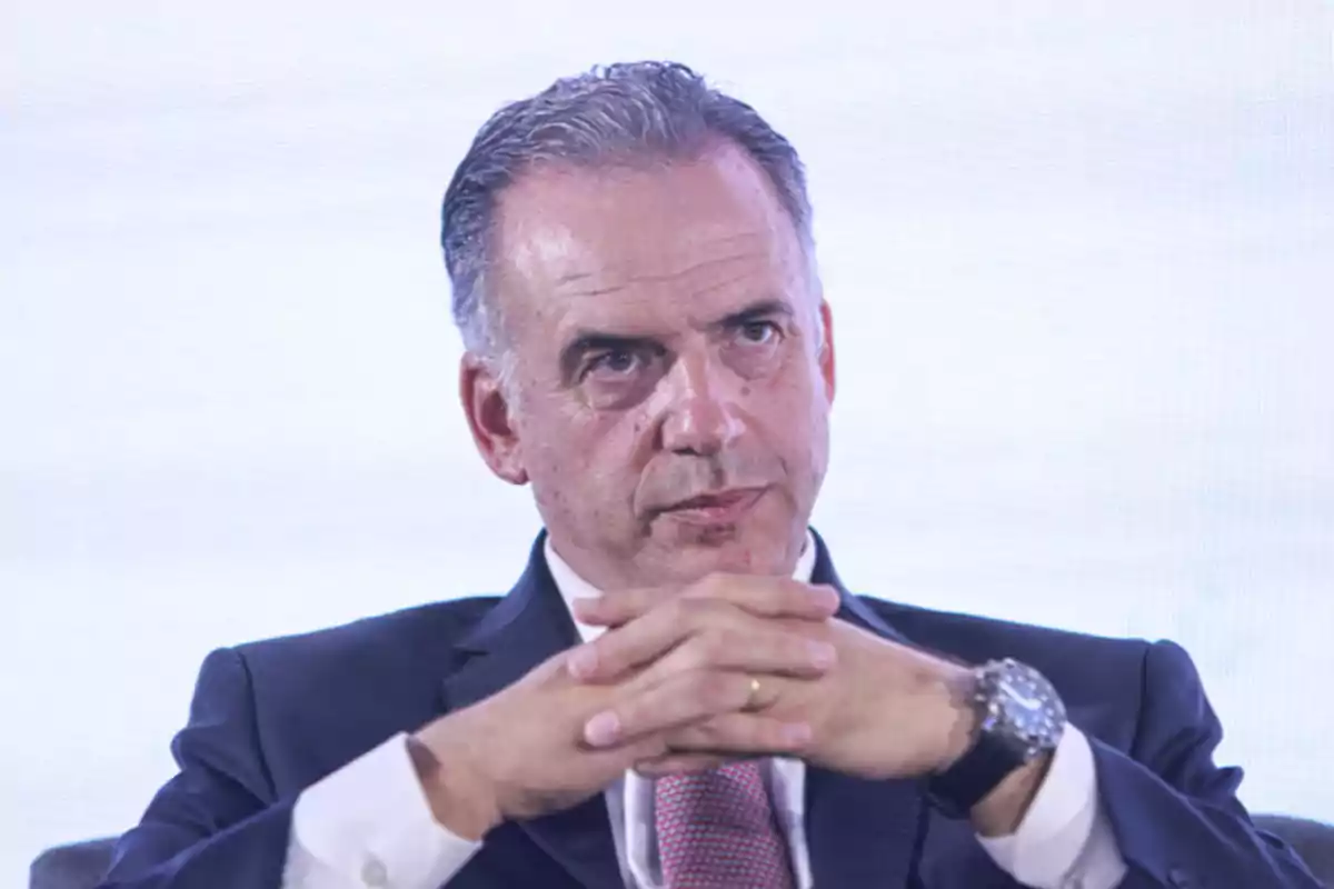 Man in a suit with a serious expression and hands clasped in front of a light background