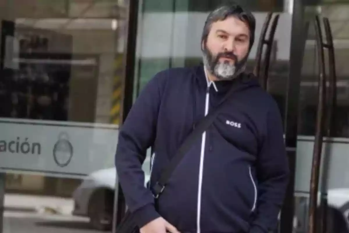 Gray-haired, bearded man wearing a blue sweatjersey with the word BOSS and carrying a black shoulder bag stands in front of a glass door.