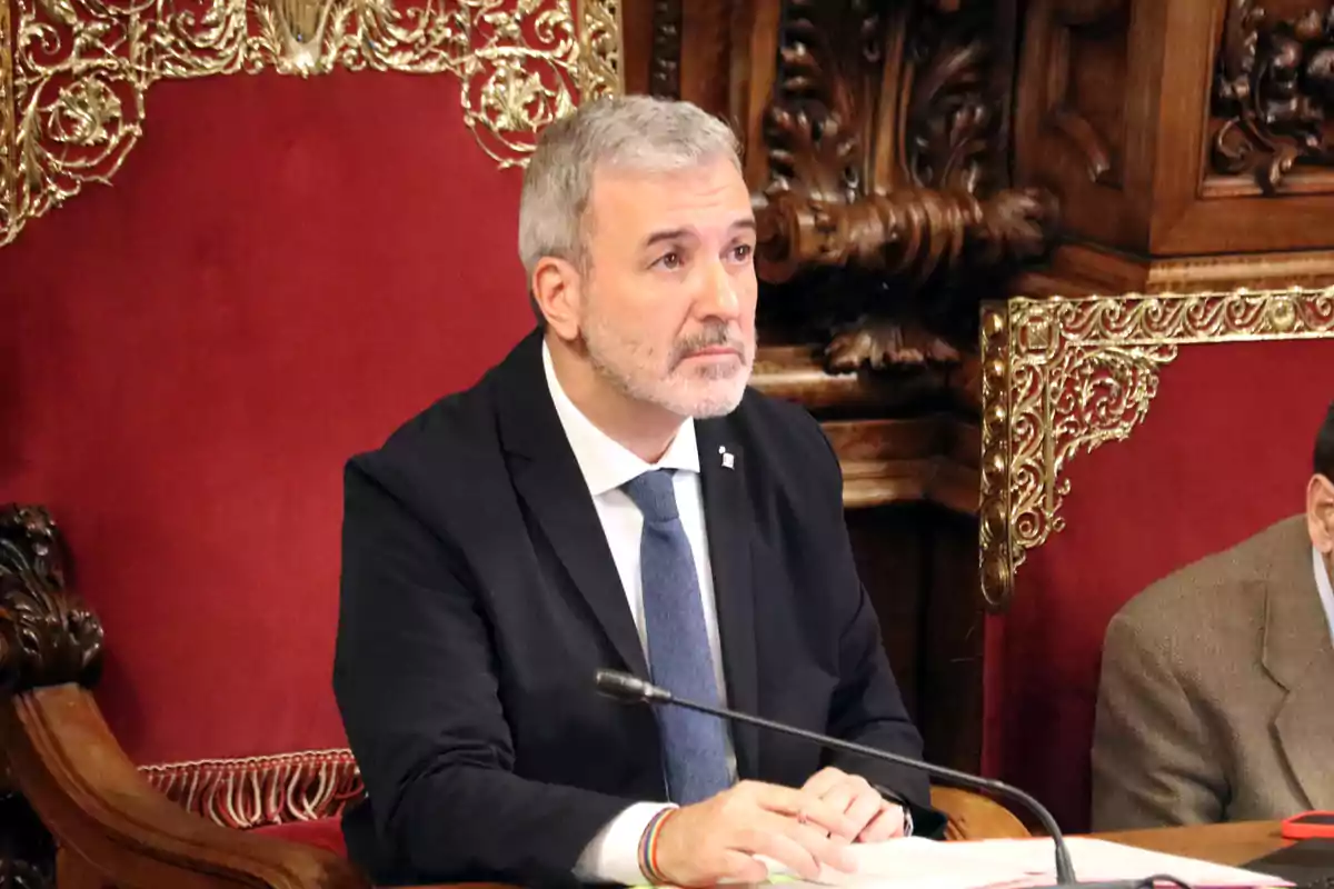 Gray-haired, bearded man sitting on a chair with a red backrest and gold details, wearing a dark suit and blue tie, in a formal setting with carved wood. Gray-haired, bearded man sitting on a chair with a red backrest and gold details, wearing a dark suit and blue tie, in a formal setting with carved wood.