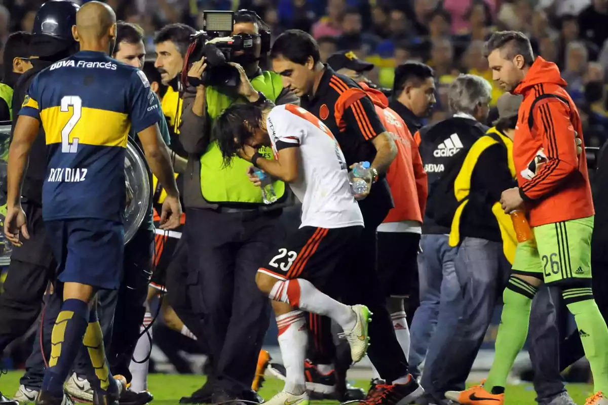A player in a white and red uniform covers his face while being assisted by team staff and surrounded by other players and cameramen in a crowded stadium. A player in a white and red uniform covers his face while being assisted by team staff and surrounded by other players and cameramen in a crowded stadium.