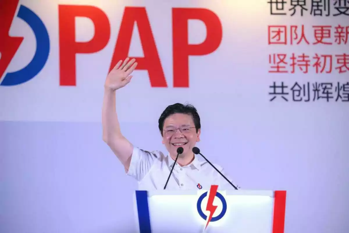 Hombre sonriente con gafas y camisa blanca saludando con la mano levantada detrás de un podio con el logo del PAP y fondo con letras en inglés y chino Hombre sonriente con gafas y camisa blanca saludando con la mano levantada detrás de un podio con el logo del PAP y fondo con letras en inglés y chino