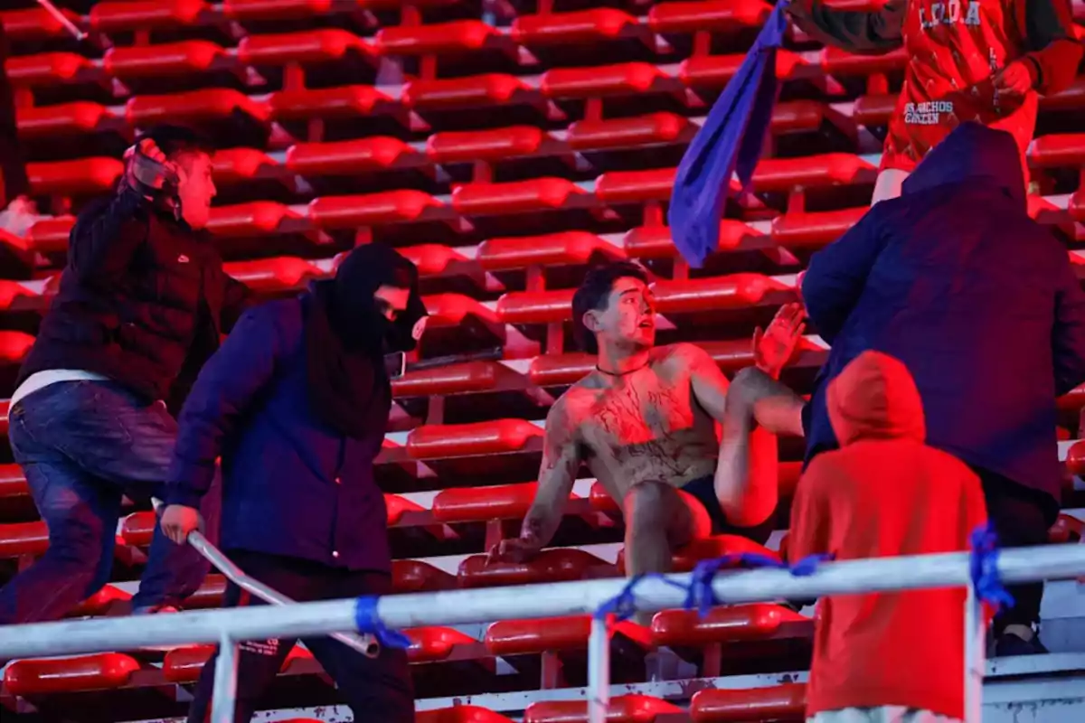 Un grupo de personas rodea a un hombre herido y semidesnudo en las gradas de un estadio iluminado con luces rojas, algunos llevan palos y el ambiente es tenso y violento Un grupo de personas rodea a un hombre herido y semidesnudo en las gradas de un estadio iluminado con luces rojas, algunos llevan palos y el ambiente es tenso y violento