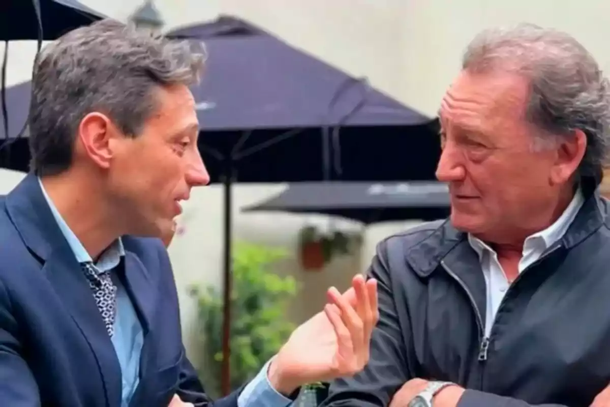 Dos hombres conversan al aire libre en un ambiente casual con sombrillas negras de fondo