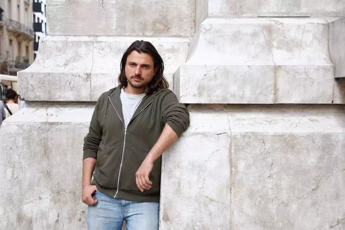 Long-haired man with a light beard leaning against a stone structure in a city Long-haired man with a light beard leaning against a stone structure in a city