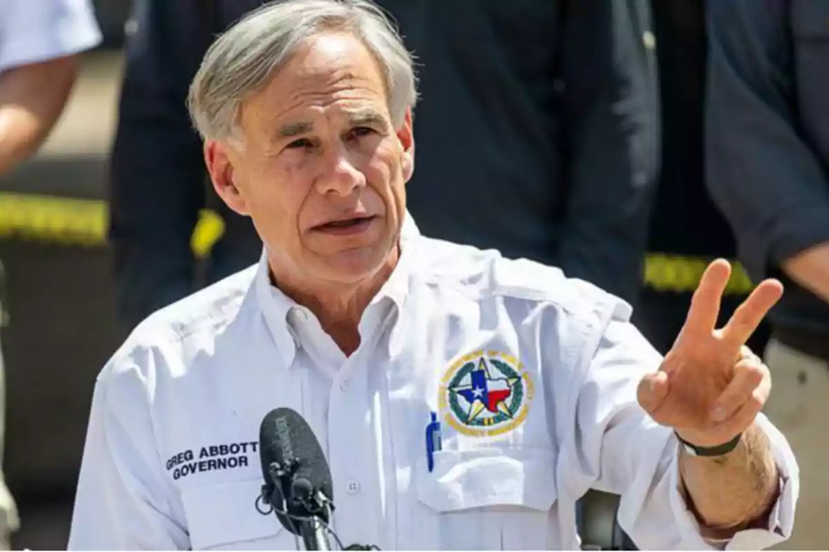 Gray-haired man in a white jersey with a microphone in front, raising two fingers of his right hand while speaking at a public event Gray-haired man in a white jersey with a microphone in front, raising two fingers of his right hand while speaking at a public event