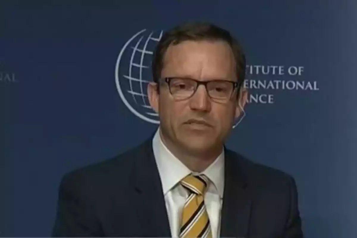 Hombre con gafas y traje oscuro hablando frente a un fondo azul con un logotipo y texto institucional Hombre con gafas y traje oscuro hablando frente a un fondo azul con un logotipo y texto institucional