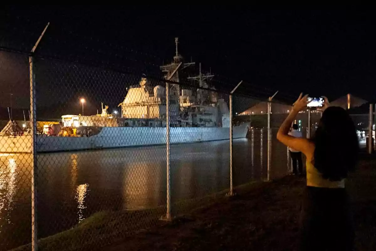 A woman takes a photo with her cellphone of a lit-up boat sailing at night behind a metal fence. A woman takes a photo with her cellphone of a lit-up boat sailing at night behind a metal fence.