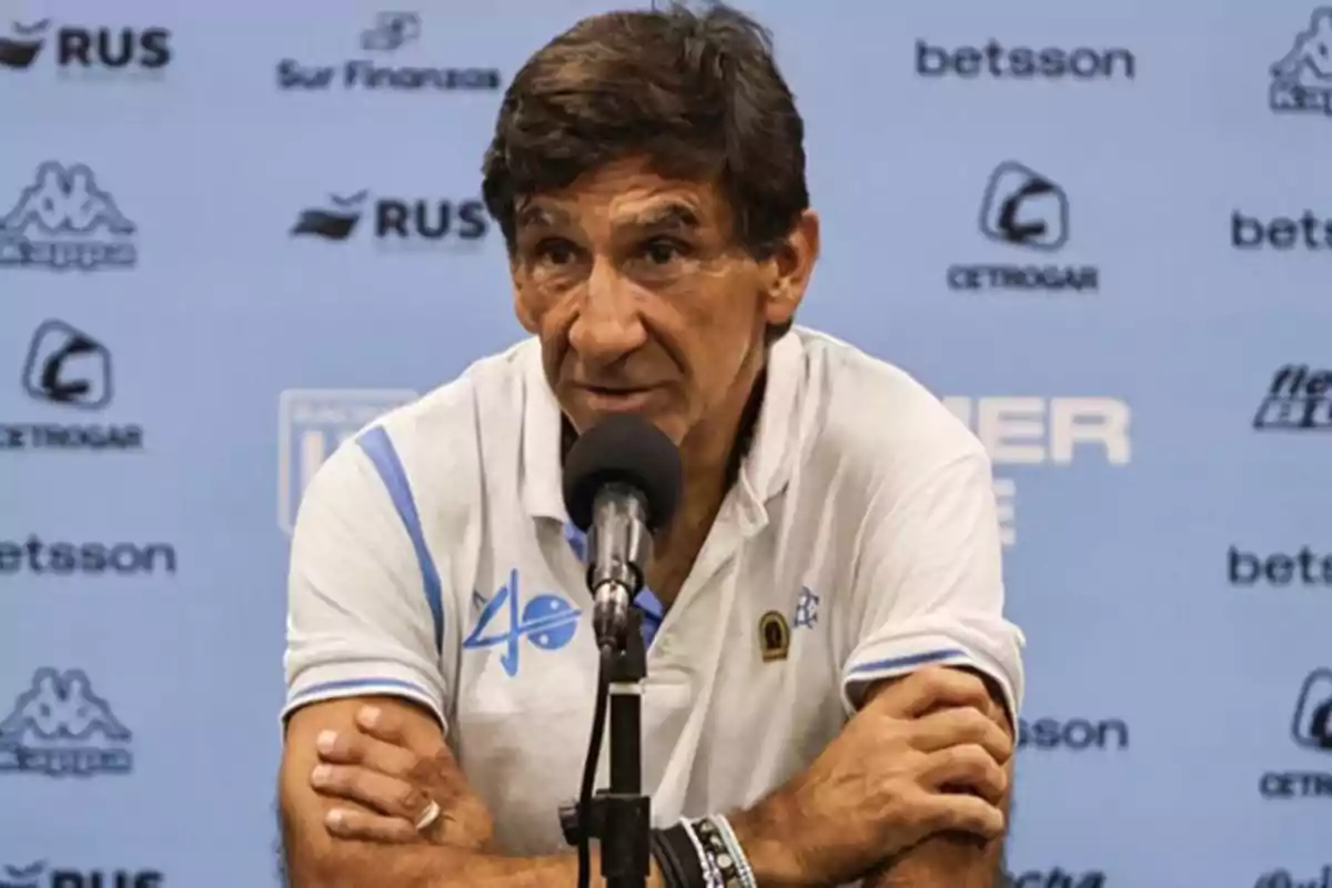 Hombre de cabello castaño y camiseta blanca con detalles celestes hablando en una conferencia de prensa frente a un micrófono con fondo azul lleno de logotipos.