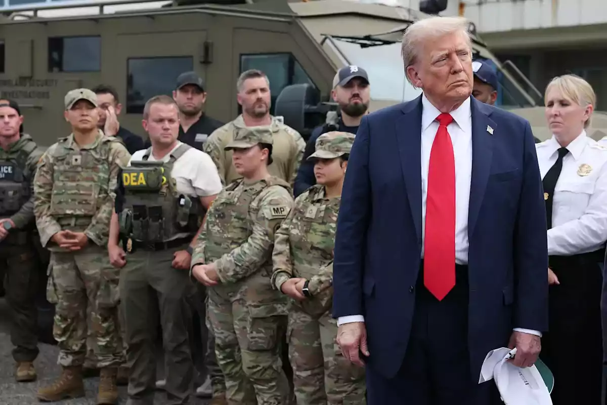 A man in a blue suit and red tie is standing in front of a group of people in military and police uniforms next to an armored vehicle.