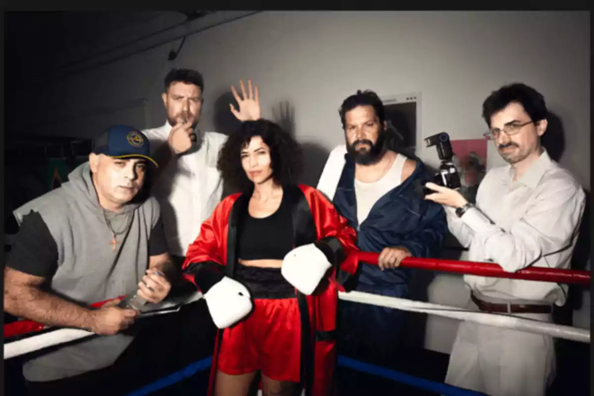 Cinco personas posan dentro de un ring de boxeo, una mujer al centro lleva guantes y ropa de boxeadora, mientras los otros cuatro hombres la rodean con diferentes expresiones y atuendos, uno de ellos sostiene una cámara fotográfica.