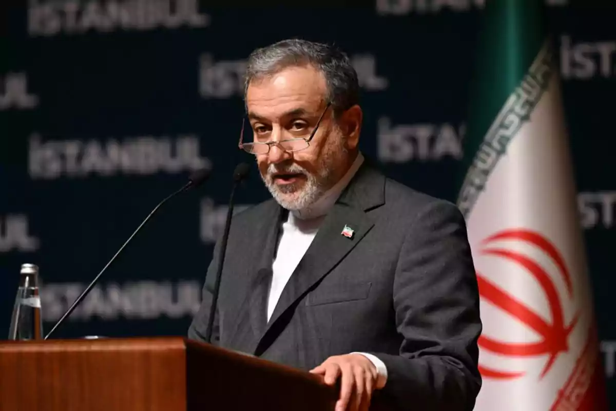 Gray-haired, bearded man speaking at a podium with microphones, wearing glasses and a gray suit, Iranian flag in the background, and the word Istanbul repeated on the dark blue backdrop. Gray-haired, bearded man speaking at a podium with microphones, wearing glasses and a gray suit, Iranian flag in the background, and the word Istanbul repeated on the dark blue backdrop.