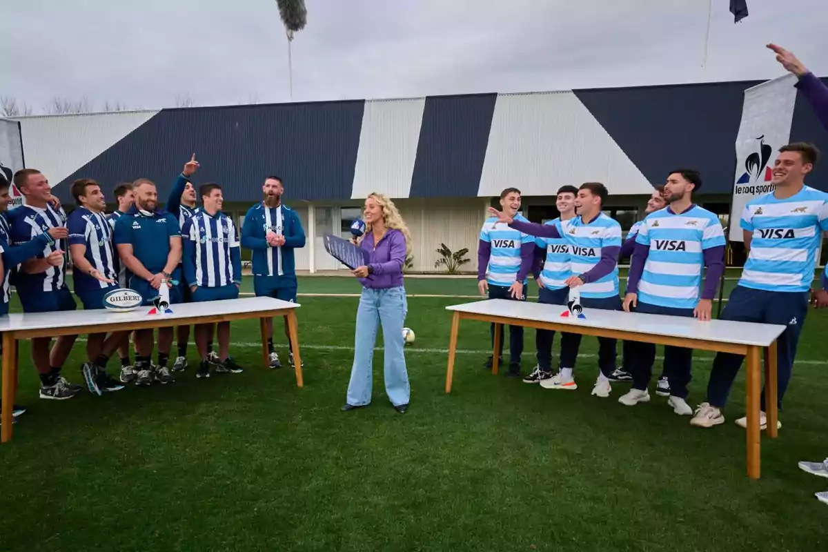 Un grupo de jugadores de rugby divididos en dos equipos, uno con camisetas a rayas azules y blancas y otro con camisetas celestes y blancas, participan en una dinámica al aire libre junto a una presentadora que sostiene un micrófono y una carpeta, todos lucen sonrientes y animados en un campo de césped frente a un edificio. Un grupo de jugadores de rugby divididos en dos equipos, uno con camisetas a rayas azules y blancas y otro con camisetas celestes y blancas, participan en una dinámica al aire libre junto a una presentadora que sostiene un micrófono y una carpeta, todos lucen sonrientes y animados en un campo de césped frente a un edificio.