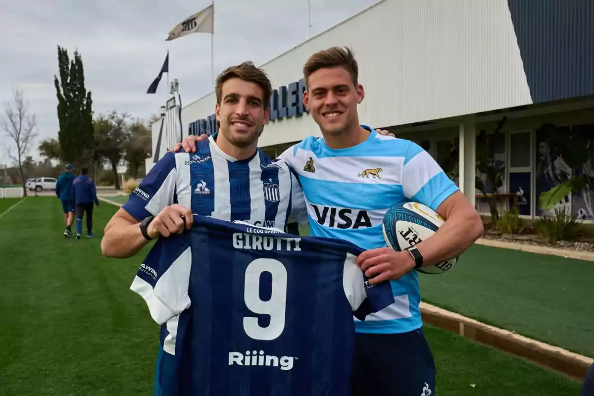 Dos deportistas sonríen y posan juntos en una cancha, uno sostiene una camiseta de fútbol con el nombre Girotti y el otro lleva una camiseta de rugby y sostiene una pelota