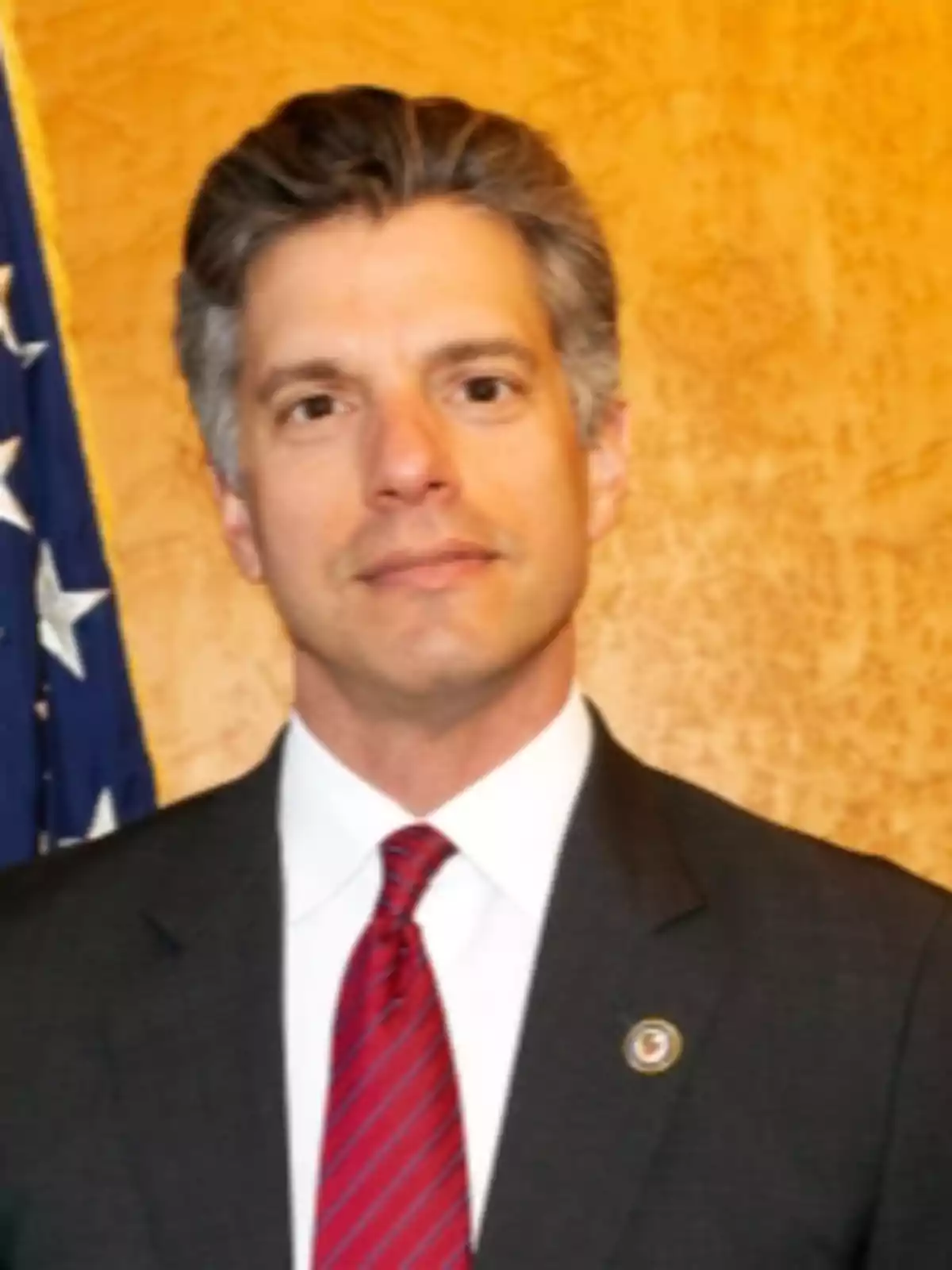 Gray-haired man in a dark suit and red tie posing in front of a yellow wall and a United States flag Gray-haired man in a dark suit and red tie posing in front of a yellow wall and a United States flag