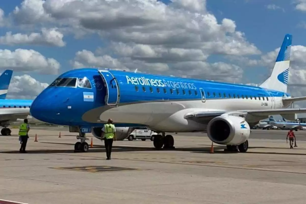 Avión de Aerolíneas Argentinas estacionado en la pista de un aeropuerto con personal de tierra alrededor y cielo parcialmente nublado de fondo Avión de Aerolíneas Argentinas estacionado en la pista de un aeropuerto con personal de tierra alrededor y cielo parcialmente nublado de fondo
