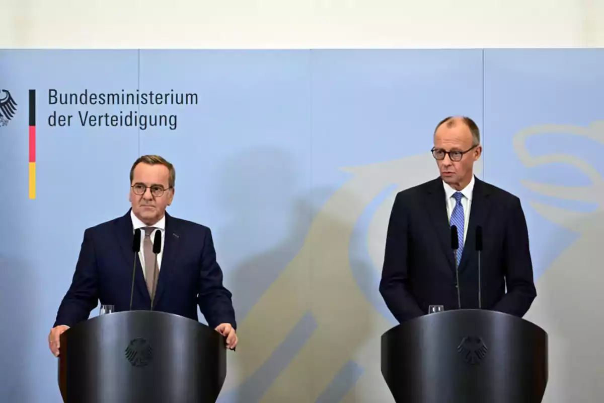Two men in suits and ties are standing behind black podiums at a press conference in front of a light blue background with the logo and name of the German Ministry of Defense. Two men in suits and ties are standing behind black podiums at a press conference in front of a light blue background with the logo and name of the German Ministry of Defense.