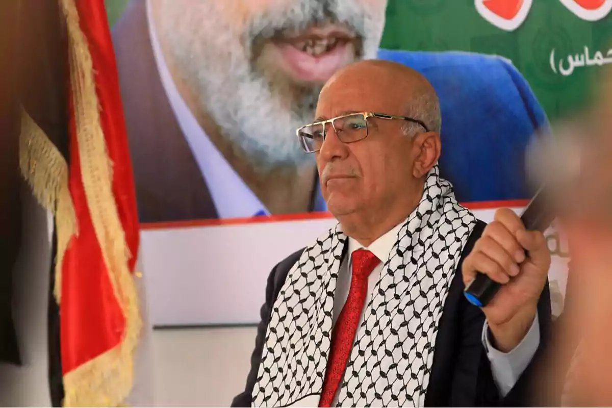 Older man with glasses and a red tie wearing a black and white keffiyeh, sitting in front of a flag and a poster with a blurred face in the background Older man with glasses and a red tie wearing a black and white keffiyeh, sitting in front of a flag and a poster with a blurred face in the background
