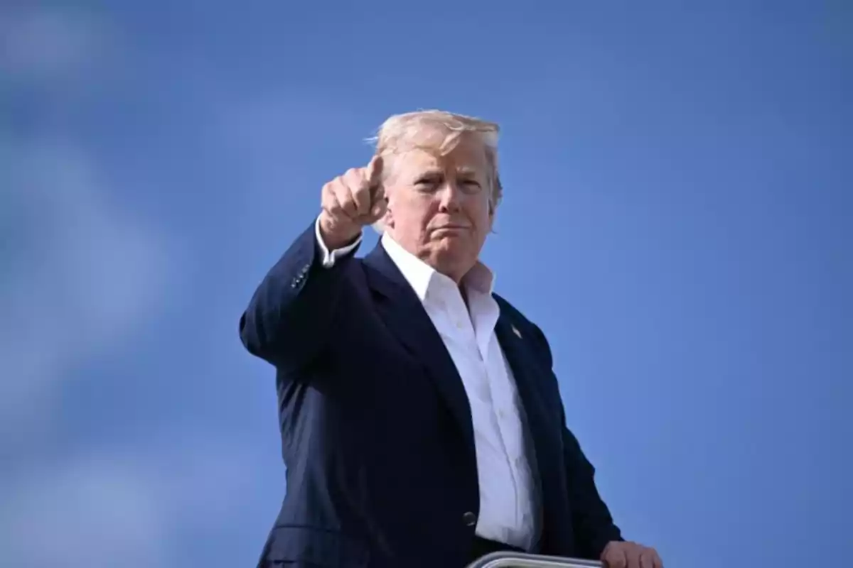 Blond-haired man in a dark suit and white jersey pointing forward with a blue sky in the background
