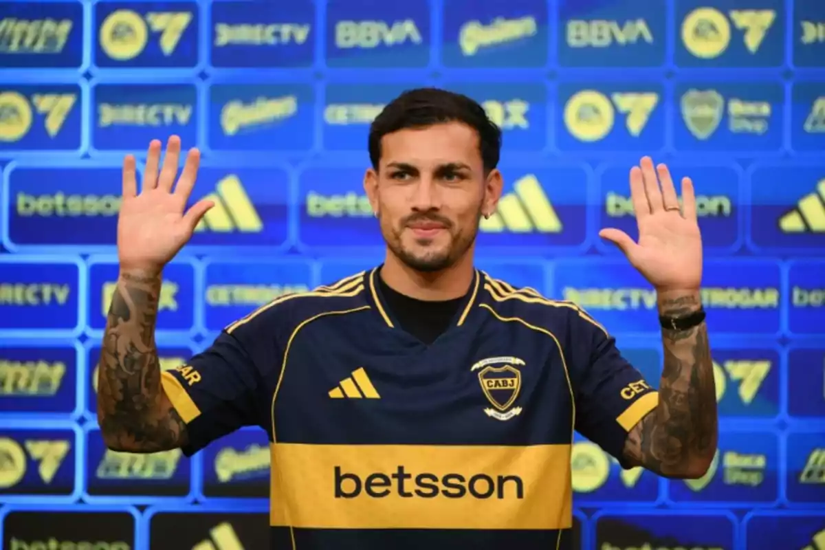 Soccer player wearing a Boca Juniors jersey raising both hands in front of a blue background with sponsor logos. Soccer player wearing a Boca Juniors jersey raising both hands in front of a blue background with sponsor logos.