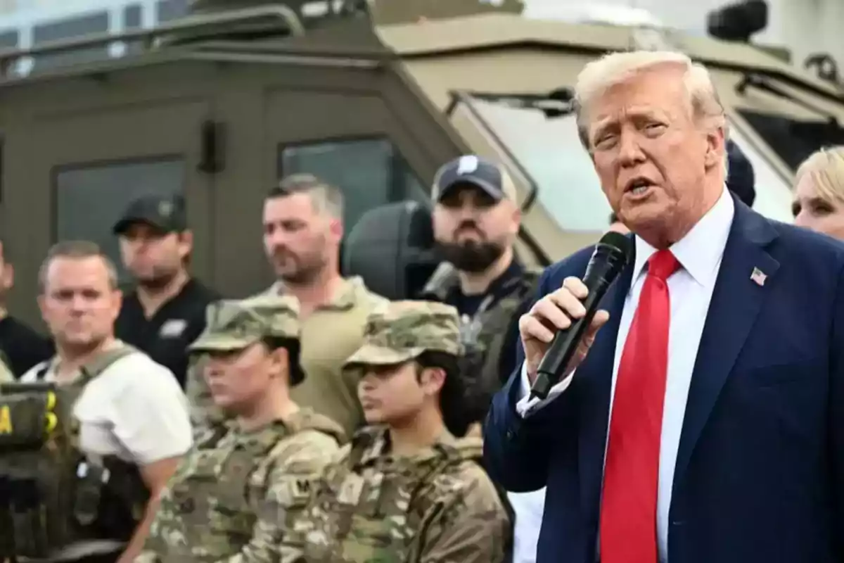 Hombre de traje azul y corbata roja hablando por micrófono frente a un grupo de personas con uniforme militar y un vehículo blindado de fondo Hombre de traje azul y corbata roja hablando por micrófono frente a un grupo de personas con uniforme militar y un vehículo blindado de fondo