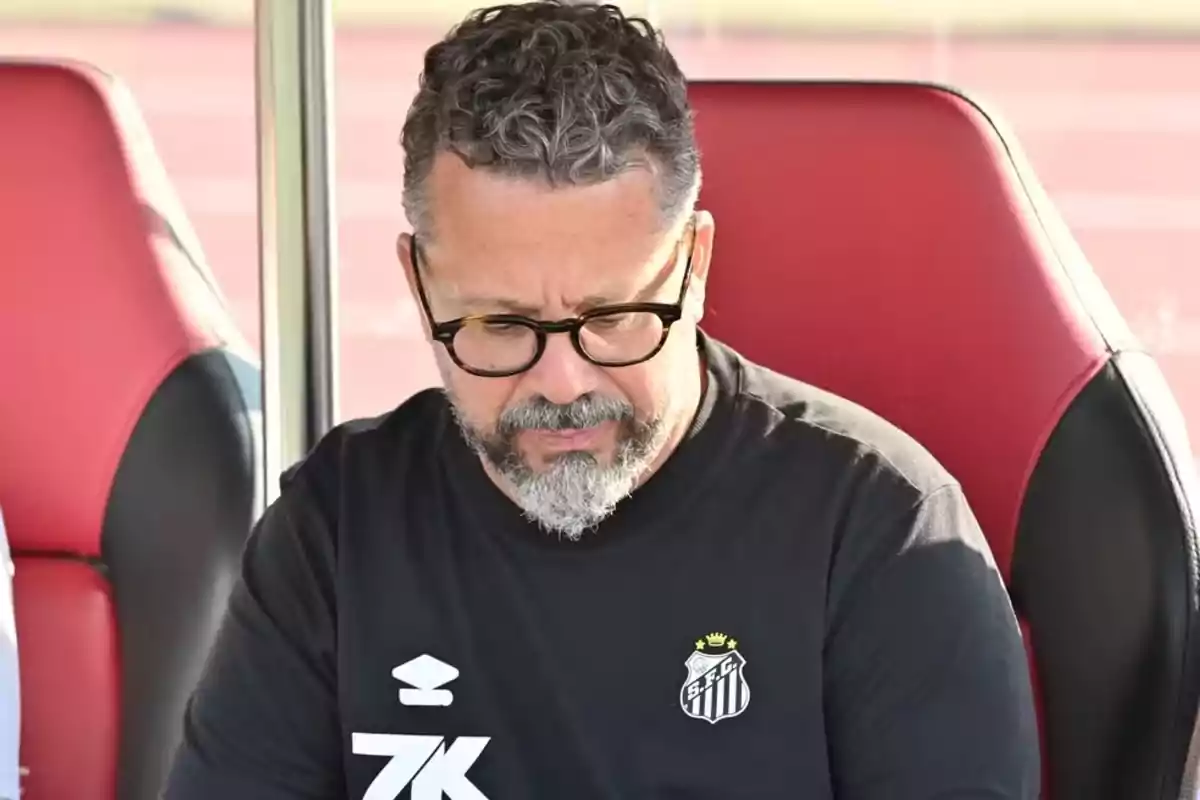 Hombre de cabello rizado y barba canosa con gafas sentado en una silla roja usando una camiseta negra del Santos FC Hombre de cabello rizado y barba canosa con gafas sentado en una silla roja usando una camiseta negra del Santos FC