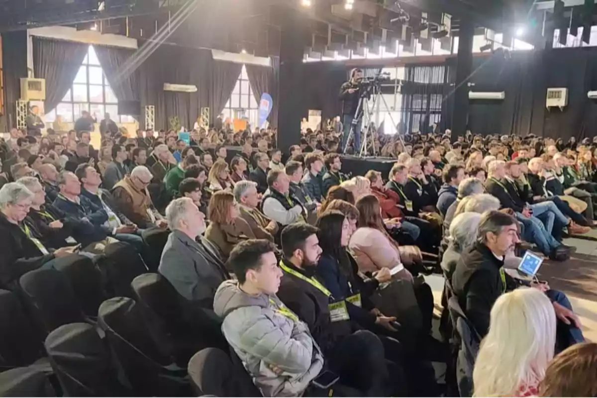 Auditorium full of seated people paying attention to an event in a brightly lit room with large windows and a cameraman filming from an elevated platform. Auditorium full of seated people paying attention to an event in a brightly lit room with large windows and a cameraman filming from an elevated platform.