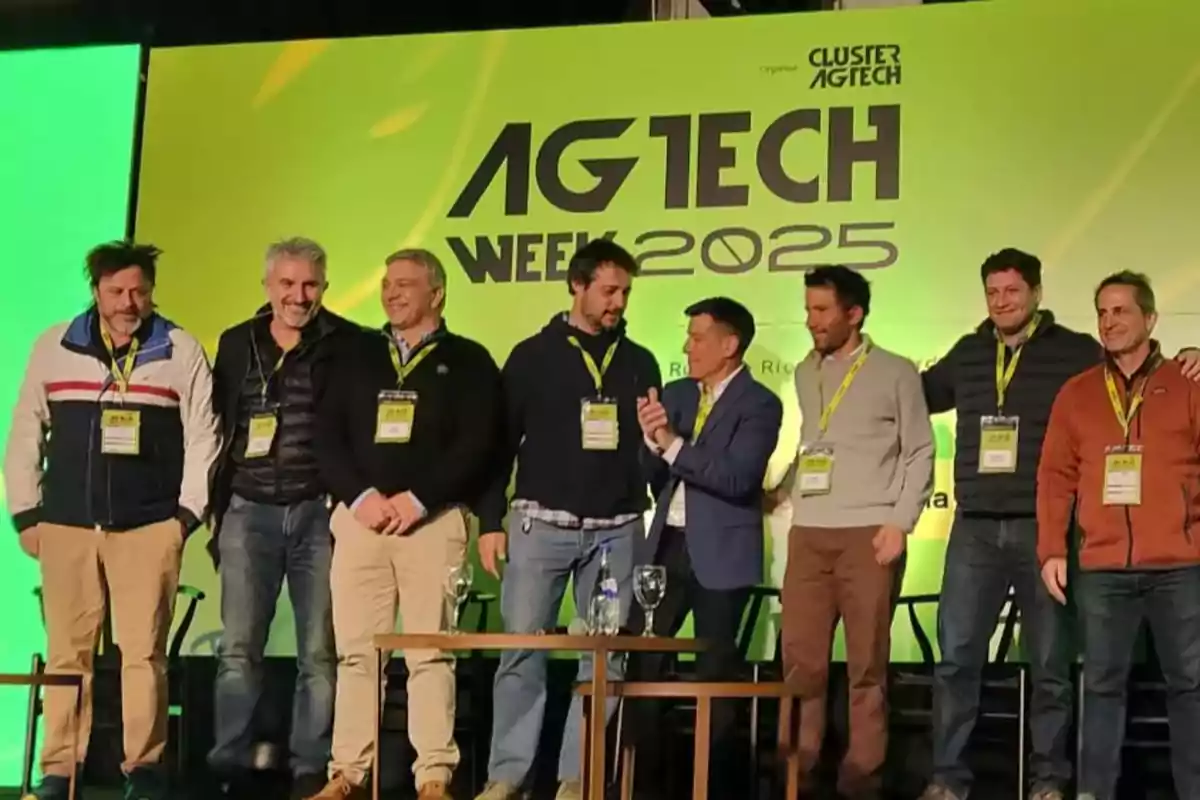 Eight men pose smiling on a stage during an agricultural technology event called AGTECH WEEK 2025 with a green and yellow background.
