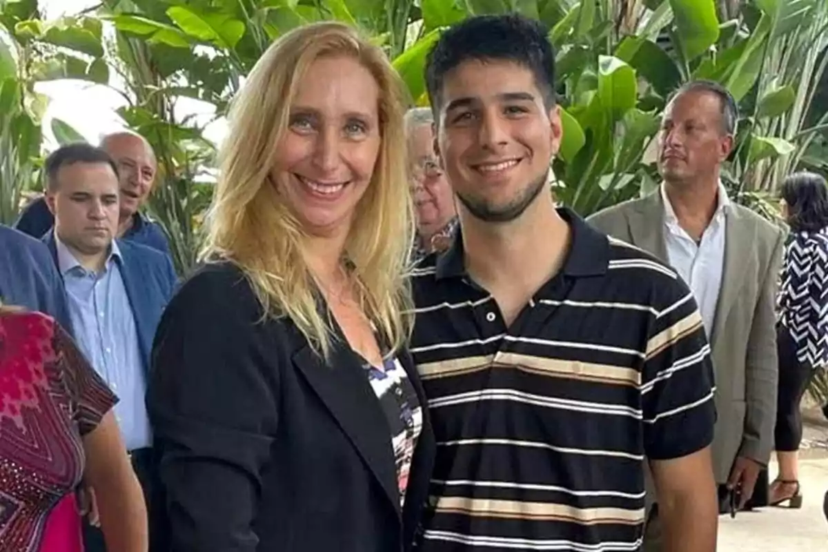 Una mujer rubia y un joven de cabello oscuro sonríen juntos en un evento al aire libre rodeados de varias personas y plantas verdes al fondo Una mujer rubia y un joven de cabello oscuro sonríen juntos en un evento al aire libre rodeados de varias personas y plantas verdes al fondo