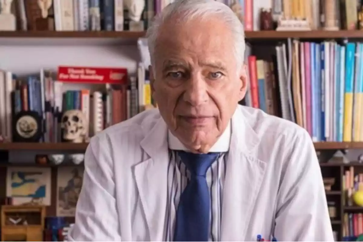 Hombre mayor con bata de médico y corbata azul frente a un estante lleno de libros