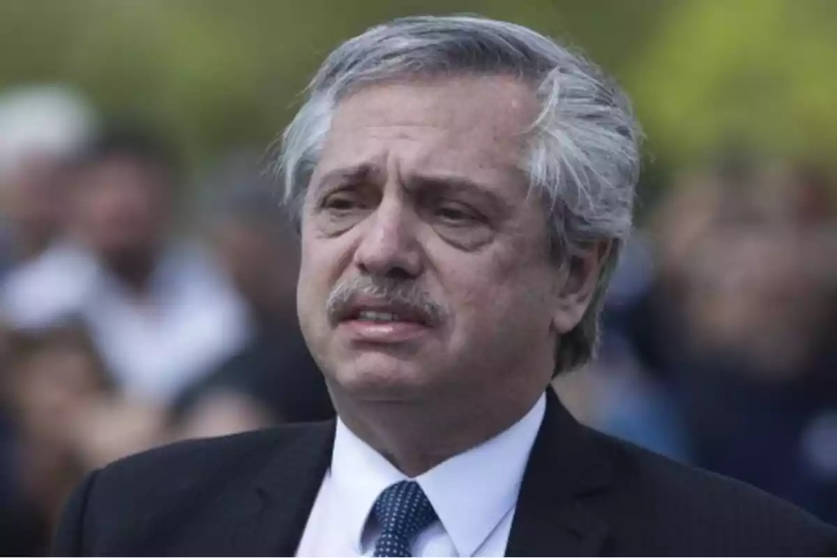 Gray-haired man with a mustache and a serious expression, wearing a suit and tie in an outdoor setting with blurred people in the background