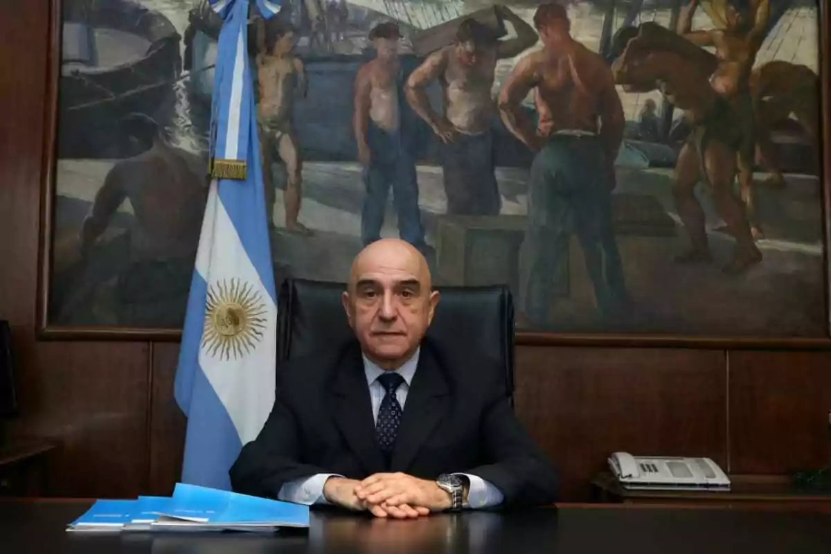 Hombre calvo de traje sentado en un escritorio con carpetas azules, una bandera de Argentina a su lado y un mural de trabajadores de fondo