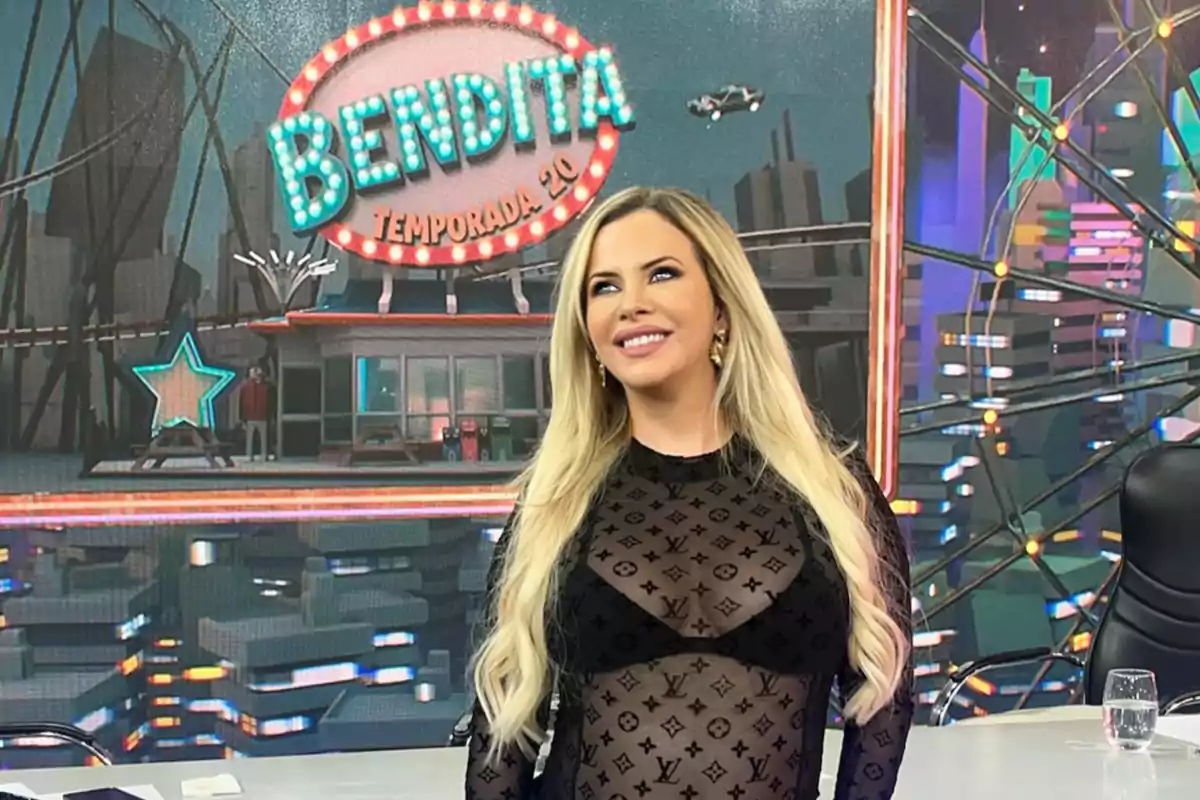 Mujer rubia con vestido negro transparente posando sonriente en un set de televisión con un cartel de Bendita Temporada 20 de fondo
