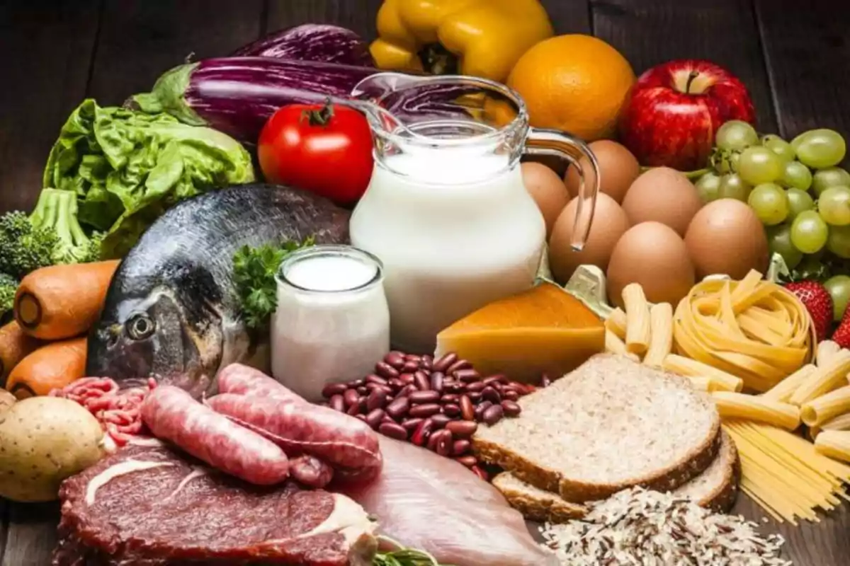 Variety of fresh foods including fruits, vegetables, bread, pasta, meat, fish, eggs, milk, cheese, yogurt, sausages, rice, and legumes on a wooden table