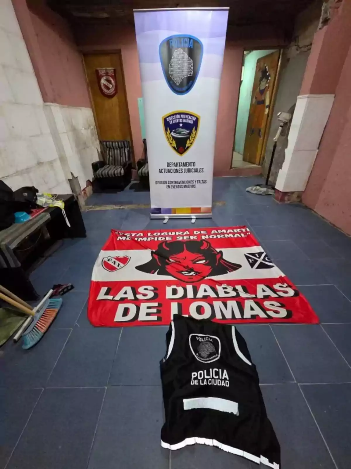 En una habitación con paredes rosadas y blancas se observa un cartel de la policía, una bandera roja con la leyenda Las Diablas de Lomas y un chaleco negro de la Policía de la Ciudad sobre el piso