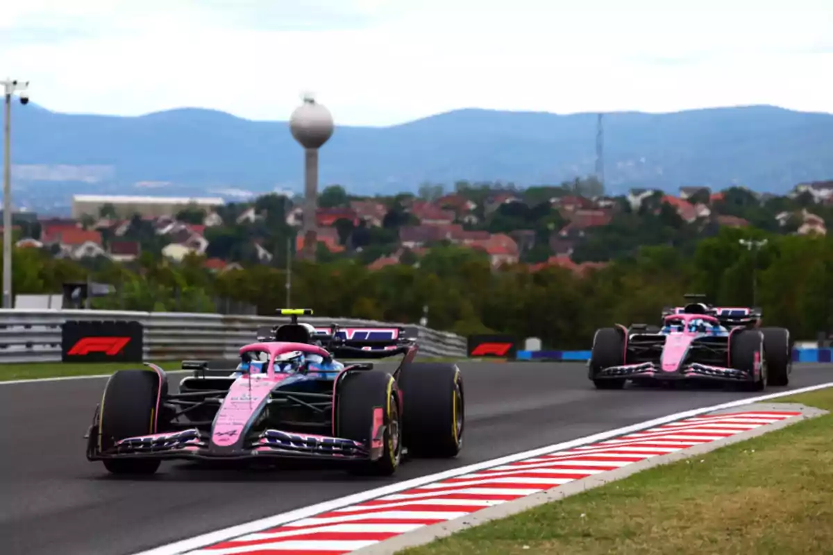 Two Formula 1 cars with pink details race on a track with a city and mountains in the background.