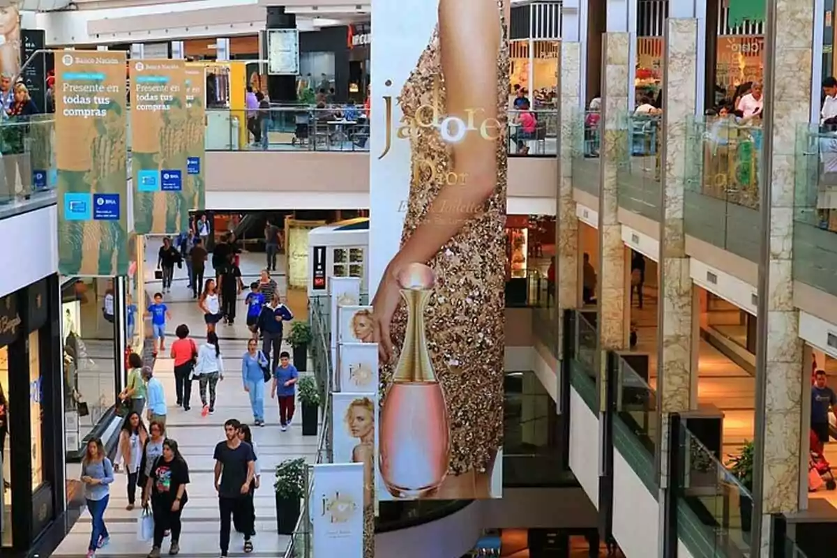 Personas caminando dentro de un centro comercial con tiendas a los lados y un gran anuncio publicitario de perfume en el centro