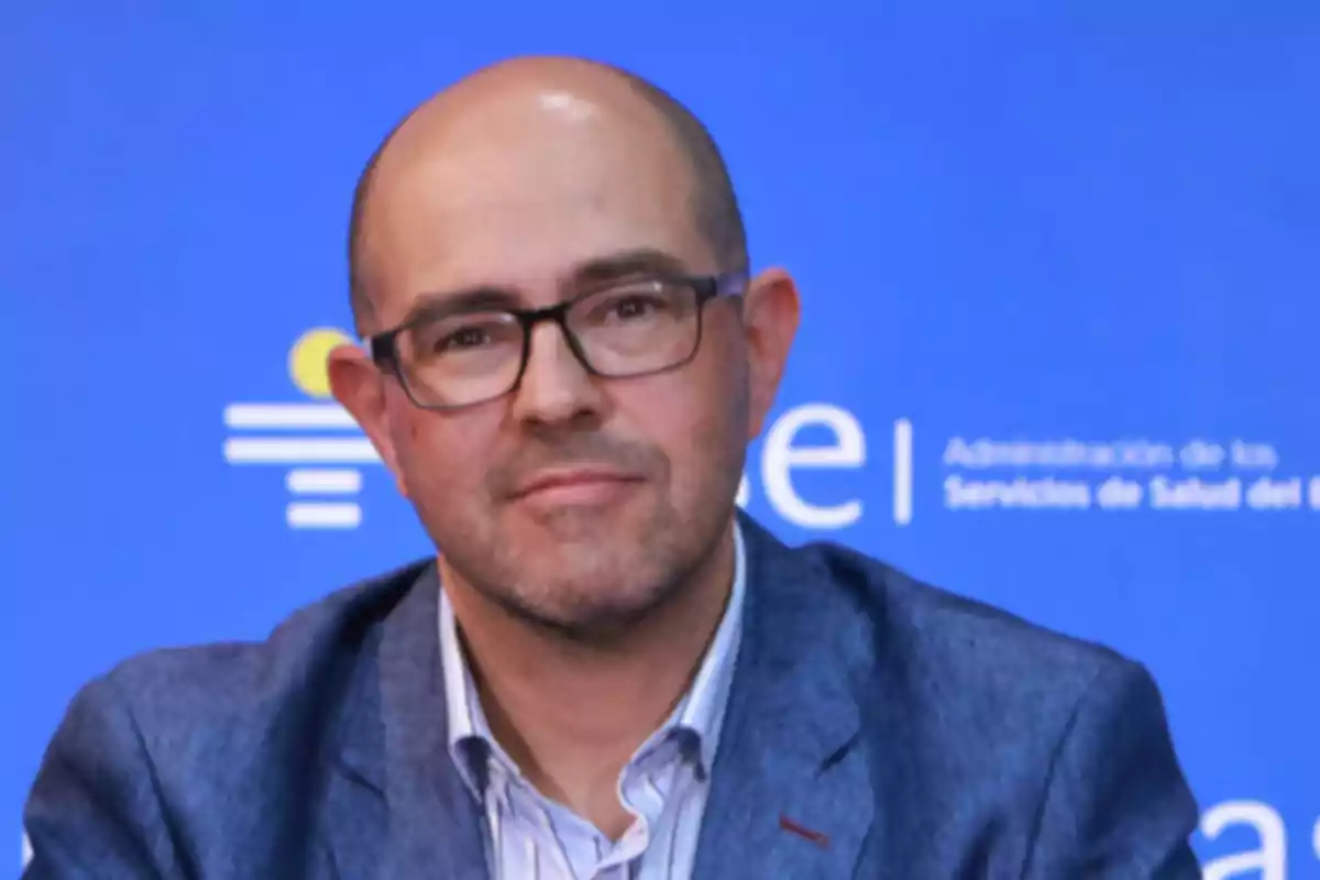 Hombre calvo con gafas y barba corta vistiendo saco azul frente a un fondo azul con texto borroso
