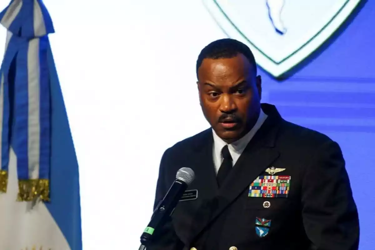 Hombre afroamericano con uniforme militar hablando frente a un micrófono junto a una bandera azul y blanca