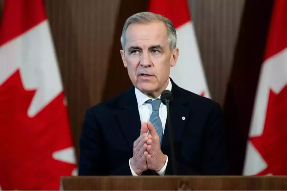 Gray-haired man in a dark suit and blue tie speaking in front of a microphone with Canadian flags in the background Gray-haired man in a dark suit and blue tie speaking in front of a microphone with Canadian flags in the background