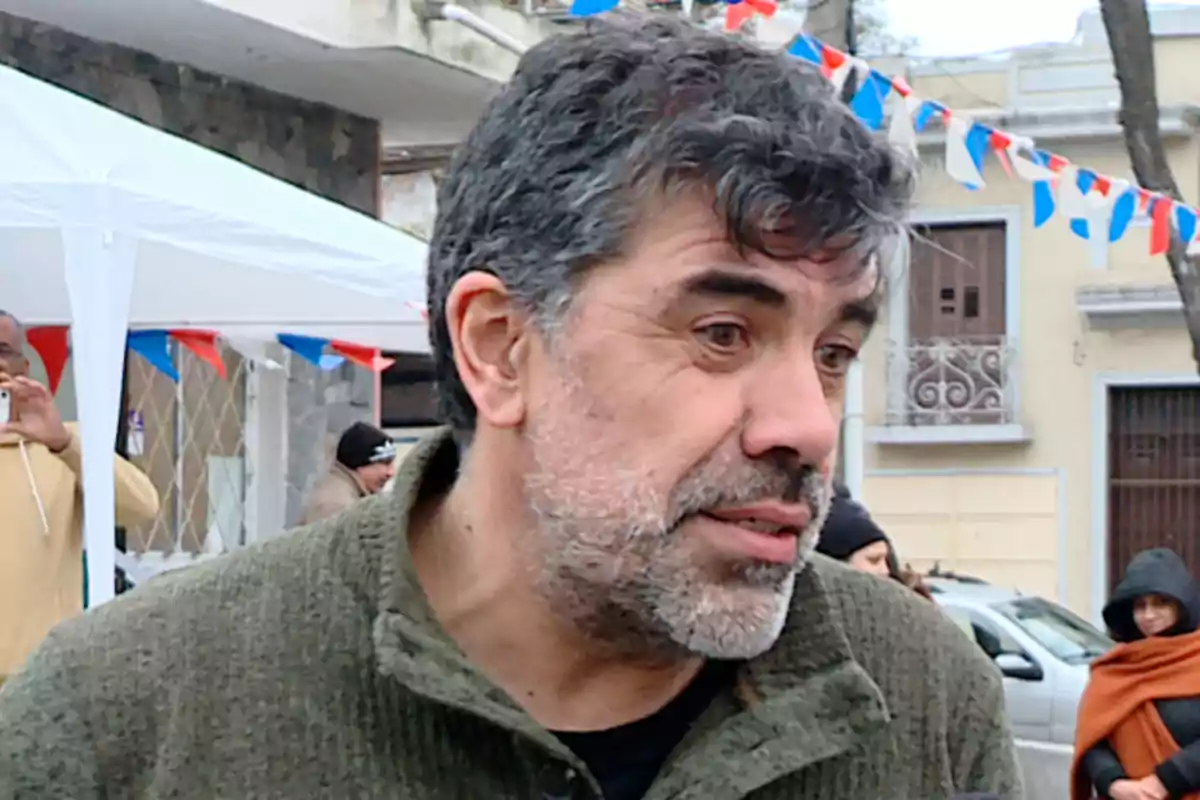 Gray-haired, bearded man speaking outdoors during an event with tricolor pennants and people in the background