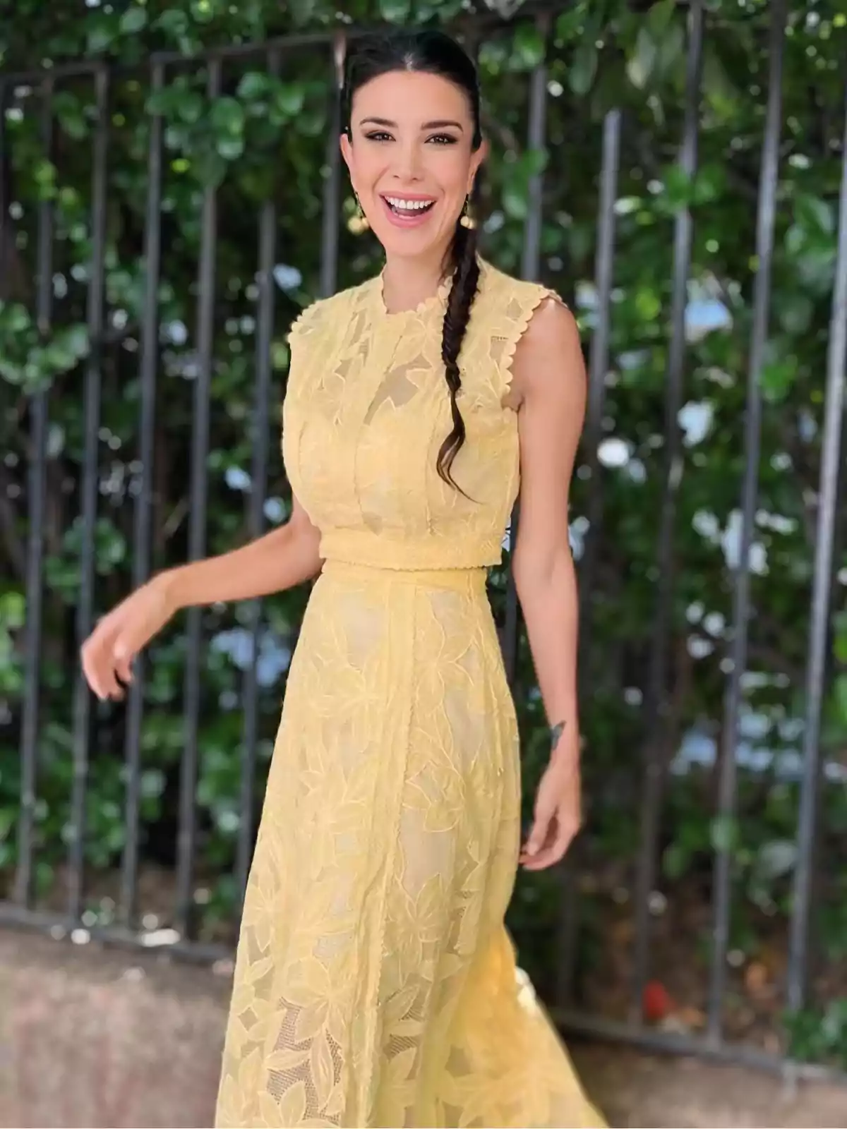 Smiling woman in a long yellow lace dress with a braid, posing outdoors in front of a fence and greenery Smiling woman in a long yellow lace dress with a braid, posing outdoors in front of a fence and greenery