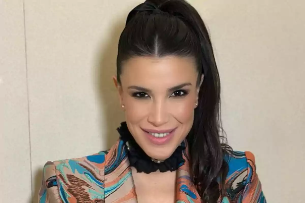 Mujer de cabello largo y oscuro recogido en una coleta alta, sonriente, con chaqueta colorida y fondo beige.