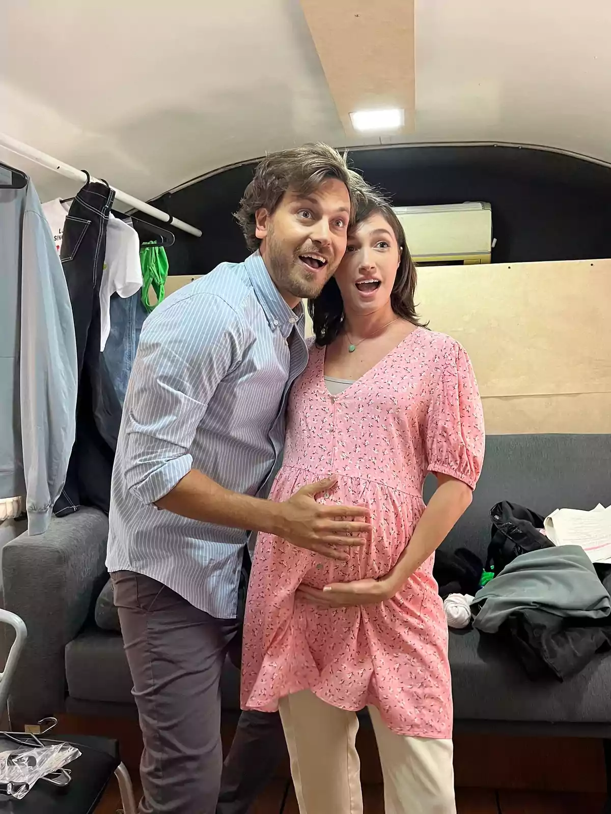 Smiling couple poses excitedly as the man holds the pregnant woman's belly in a room with clothes and objects in the background. Smiling couple poses excitedly as the man holds the pregnant woman's belly in a room with clothes and objects in the background.