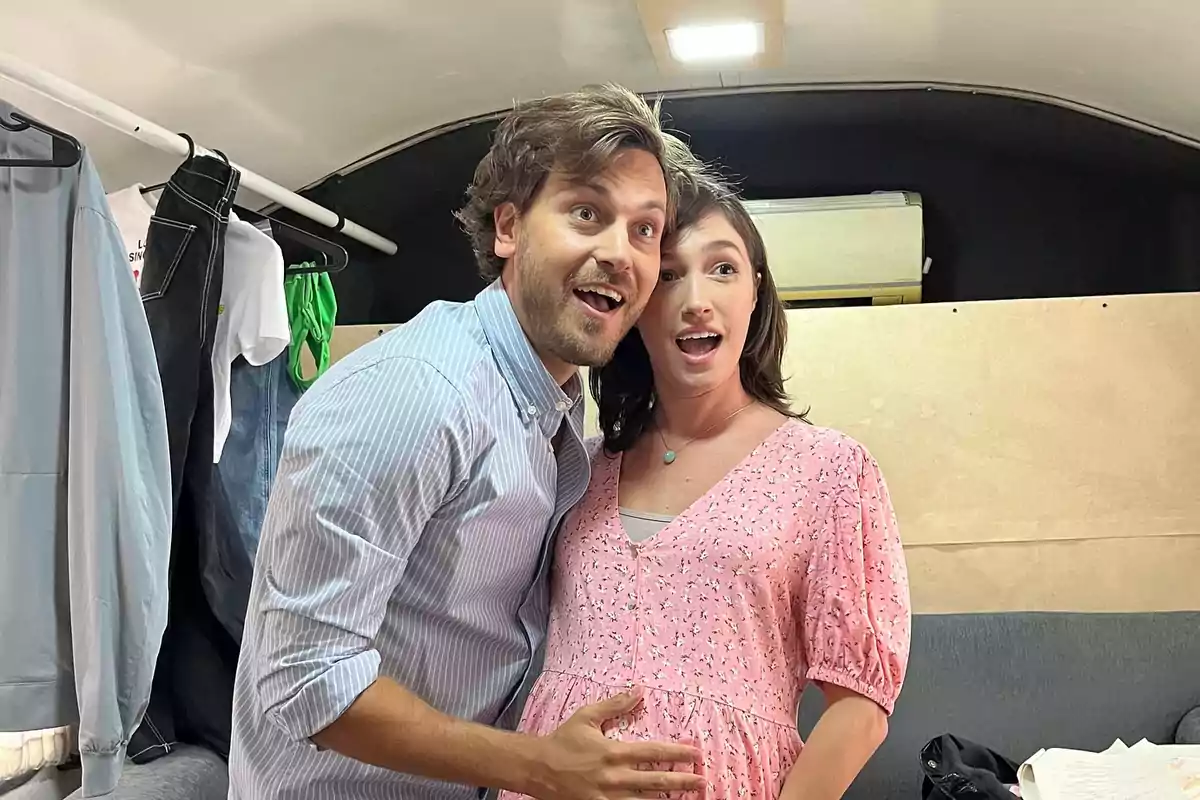 Pareja sonriente con expresión de sorpresa mientras el hombre toca la barriga de la mujer que lleva un vestido rosa en una habitación con ropa colgada al fondo