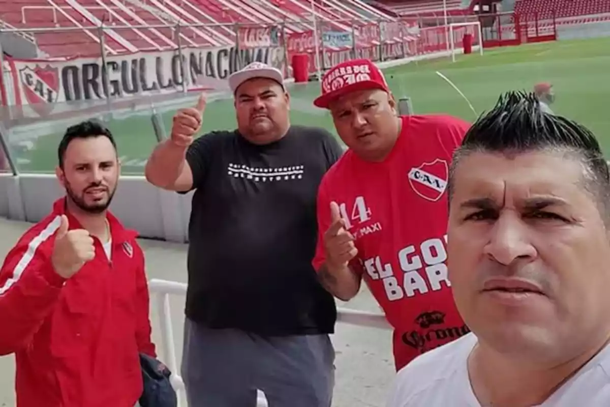 Cuatro hombres posan mostrando el pulgar arriba en un estadio de fútbol con gradas y banderas rojas de fondo Cuatro hombres posan mostrando el pulgar arriba en un estadio de fútbol con gradas y banderas rojas de fondo