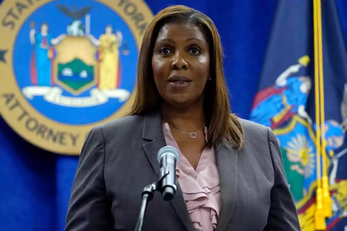 Woman with straight brown hair wearing a gray blazer and pink blouse speaking in front of a microphone with an official emblem and a flag in the background Woman with straight brown hair wearing a gray blazer and pink blouse speaking in front of a microphone with an official emblem and a flag in the background