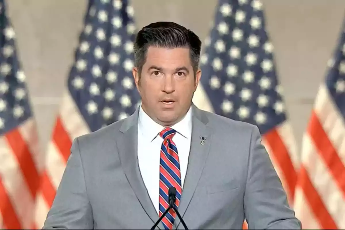 Dark-haired man in a gray suit and a red-and-blue striped tie speaking in front of several blurred United States flags in the background. Dark-haired man in a gray suit and a red-and-blue striped tie speaking in front of several blurred United States flags in the background.