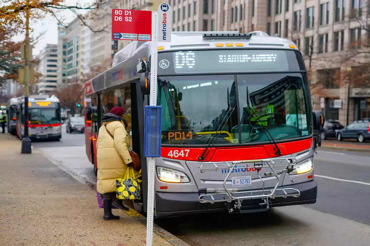 Persona con abrigo amarillo subiendo a un autobús urbano en una ciudad con edificios altos al fondo Persona con abrigo amarillo subiendo a un autobús urbano en una ciudad con edificios altos al fondo