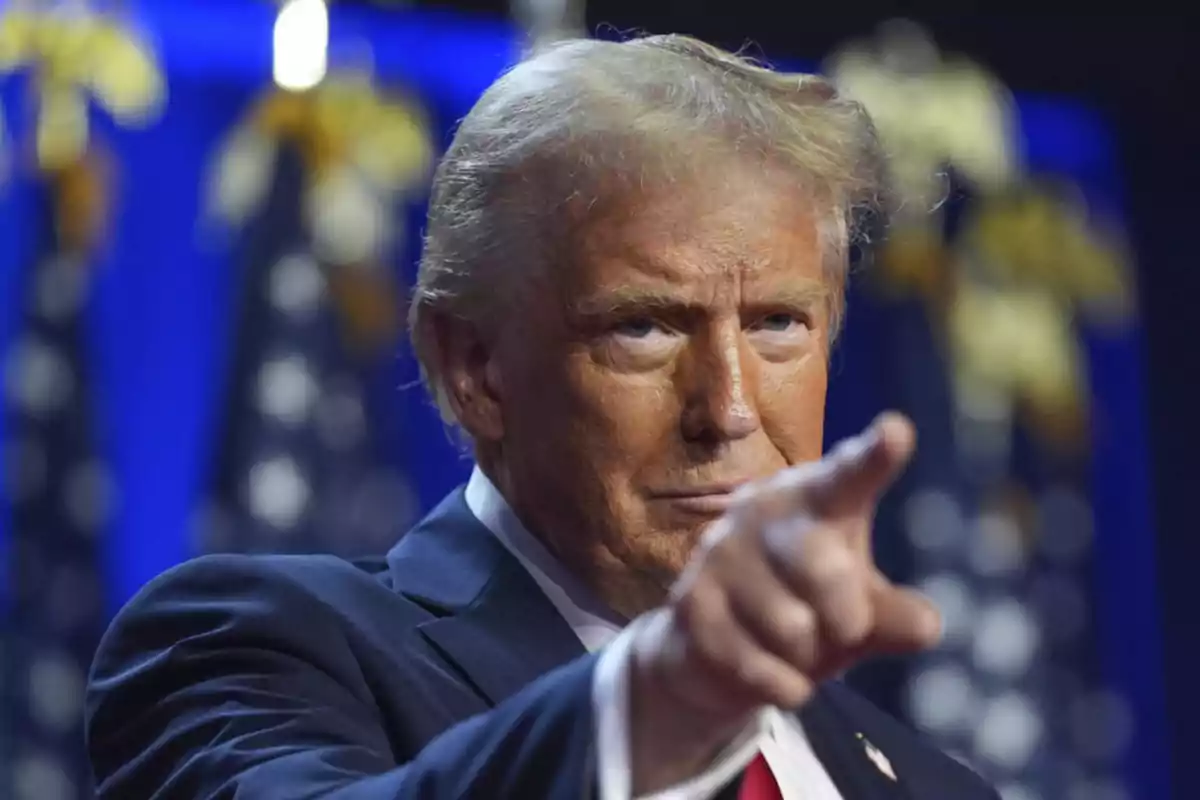 Blond-haired man in a blue suit pointing forward with a serious expression and blurred flags in the background Blond-haired man in a blue suit pointing forward with a serious expression and blurred flags in the background